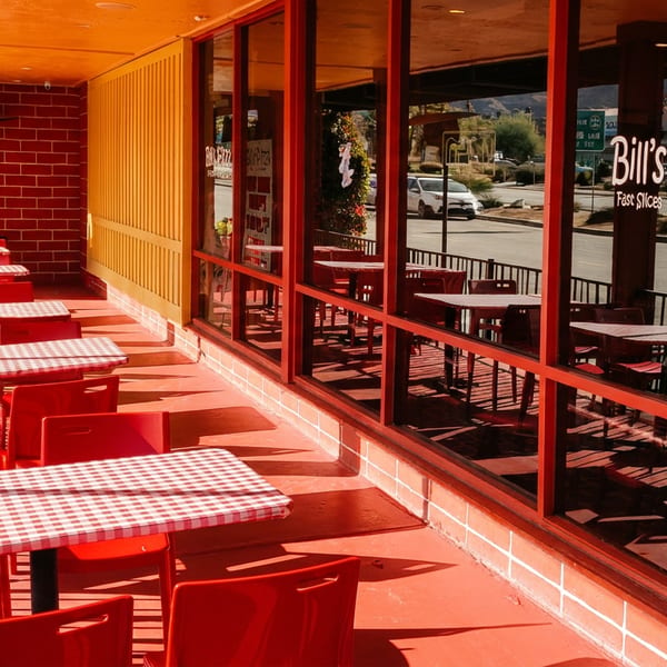 Outdoor patio of Bill's Pizza with red chairs, checkered tables, and large windows reflecting the street.
