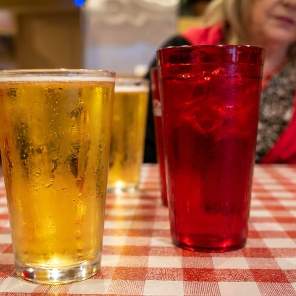 Two glasses of beer and a red drink on a red and white checkered tablecloth.