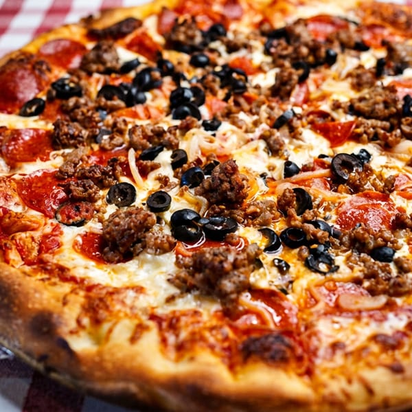 A whole pizza with pepperoni, sausage, black olives, and onions on a red and white checkered tablecloth.