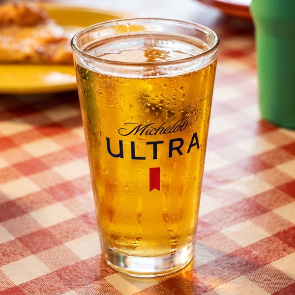 A Michelob Ultra beer in a glass on a red and white checkered tablecloth with pizza.