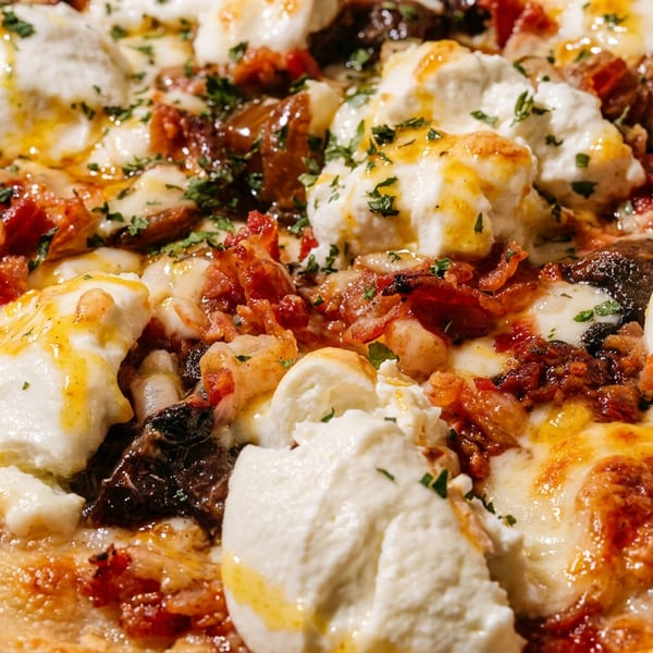Close-up of a pizza with melted cheese, burrata, crispy bacon, and fresh herbs drizzled with oil.