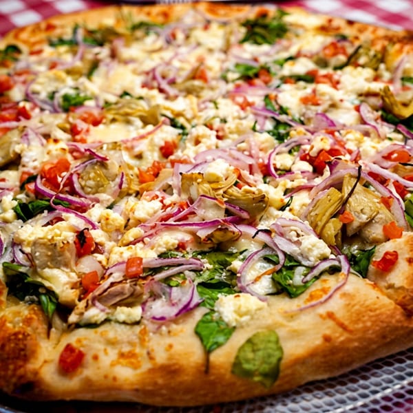 A whole pizza with spinach, artichoke hearts, red onions, tomatoes, and feta cheese on a red and white checkered tablecloth.