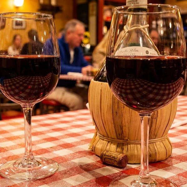Two glasses of red wine and a straw-covered bottle on a checkered tablecloth in a cozy restaurant.