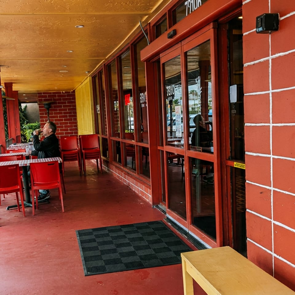 A man sips a drink at an outdoor patio table with red chairs and a red floor.