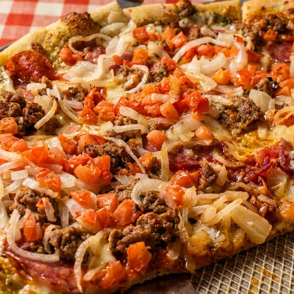 Close-up of a pizza with sausage, pepperoni, onions, and diced tomatoes on a red and white checkered tablecloth.