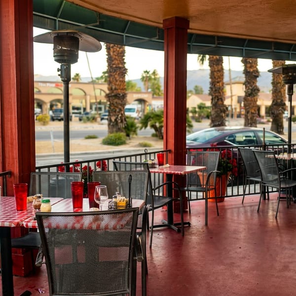 Outdoor patio dining area with red and white checkered tables, metal chairs, and palm trees in the background.