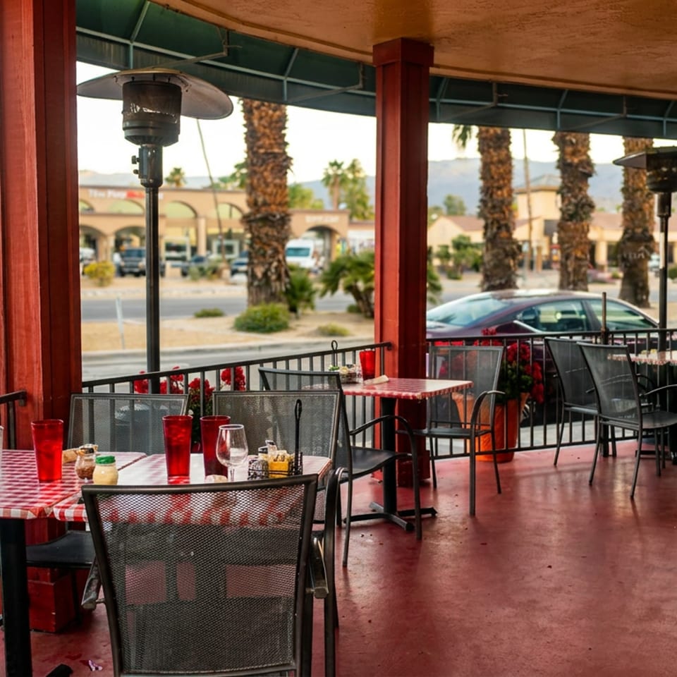Outdoor patio dining area with red and white checkered tables, metal chairs, and palm trees in the background.