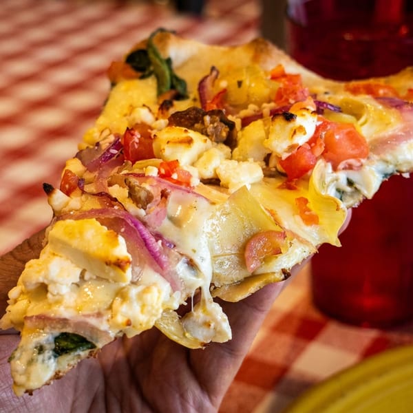 A hand holds a gourmet pizza slice with feta, red onions, tomatoes, and artichoke hearts.