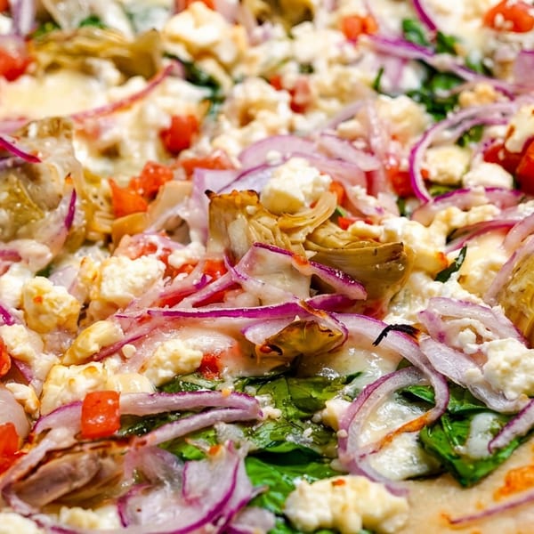 Close-up of a pizza topped with red onions, feta cheese, artichoke hearts, spinach, and diced tomatoes.