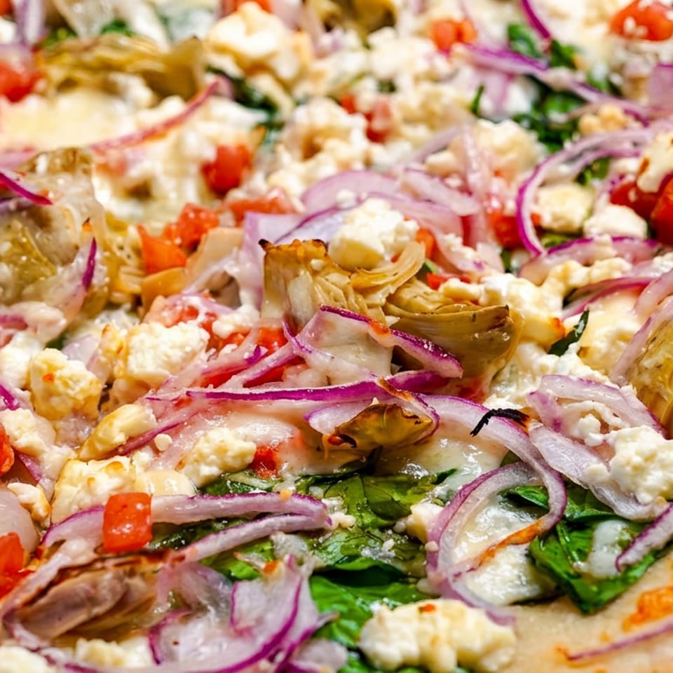 Close-up of a pizza topped with red onions, feta cheese, artichoke hearts, spinach, and diced tomatoes.