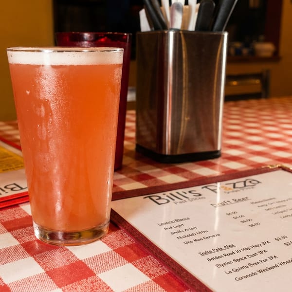 A frosty glass of pink beer sits on a red and white checkered tablecloth next to a Bill's Pizza menu.